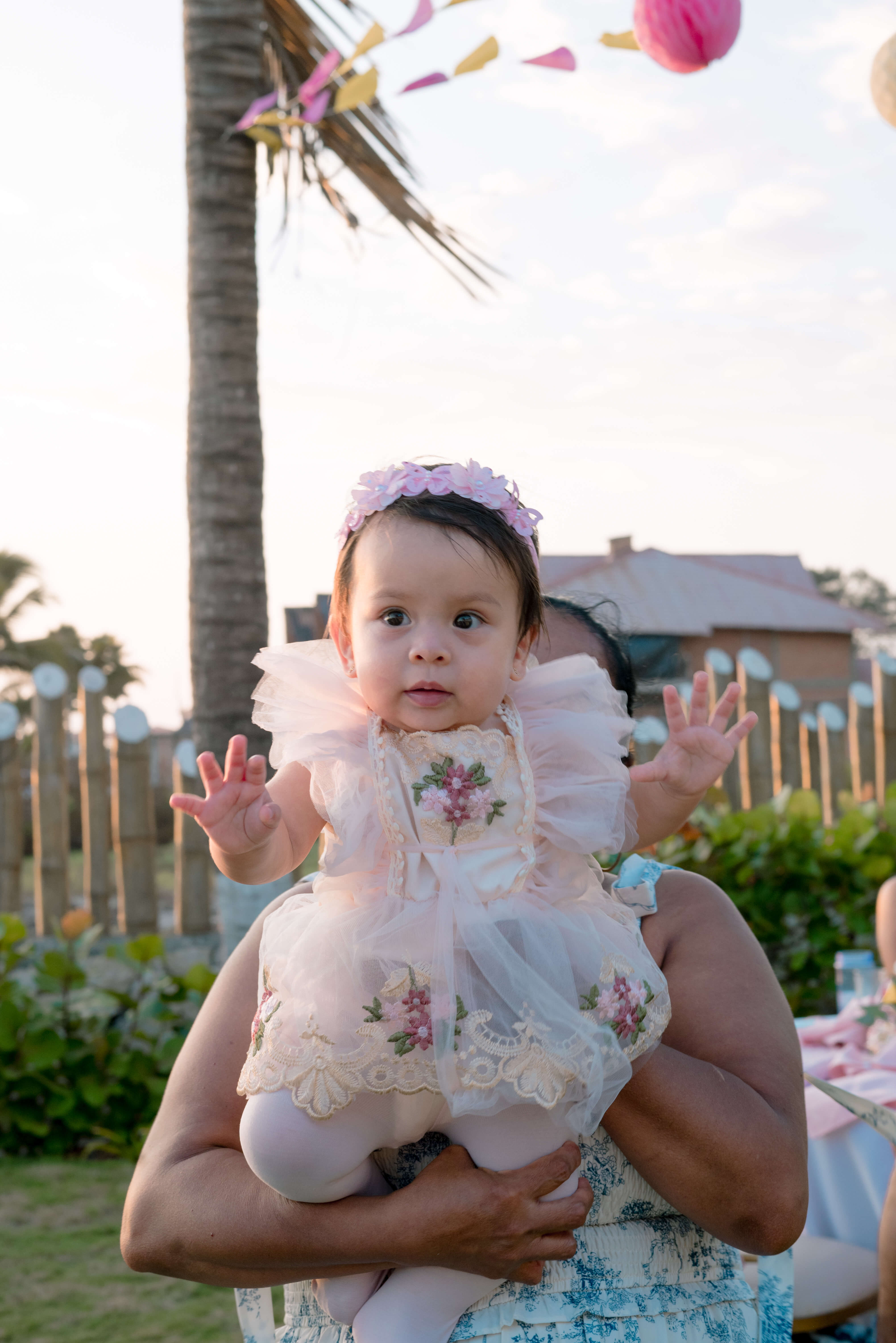 Bebé Mía con vestido de encaje floral y diadema rosada en brazos durante su matiné de primer año en Las Marías Playas
